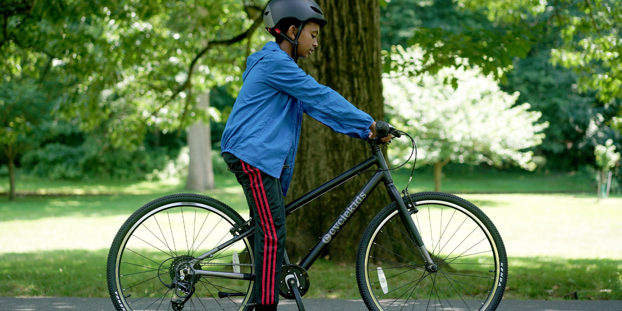 Pre-teen boy standing with his 26" CYCLEKids bike in black with a matching helmet on a path at the park
